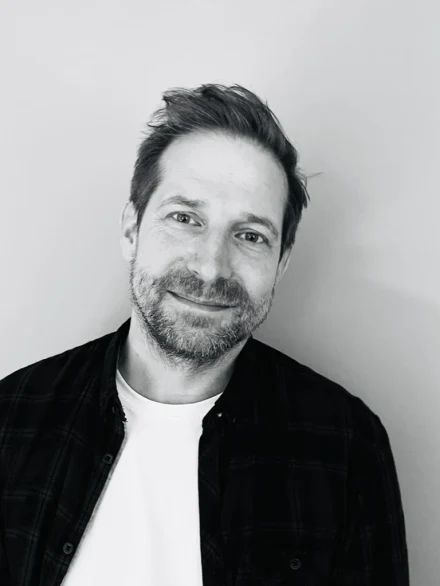Hendrik Eilers, Schwarzweißaufnahme vor einer hellen Wand: kurzes dunkles Haar, Dreitagebart, dunkel kariertes Hemd über weißem T-Shirt. Er sieht in die Kamera und lächelt leicht.